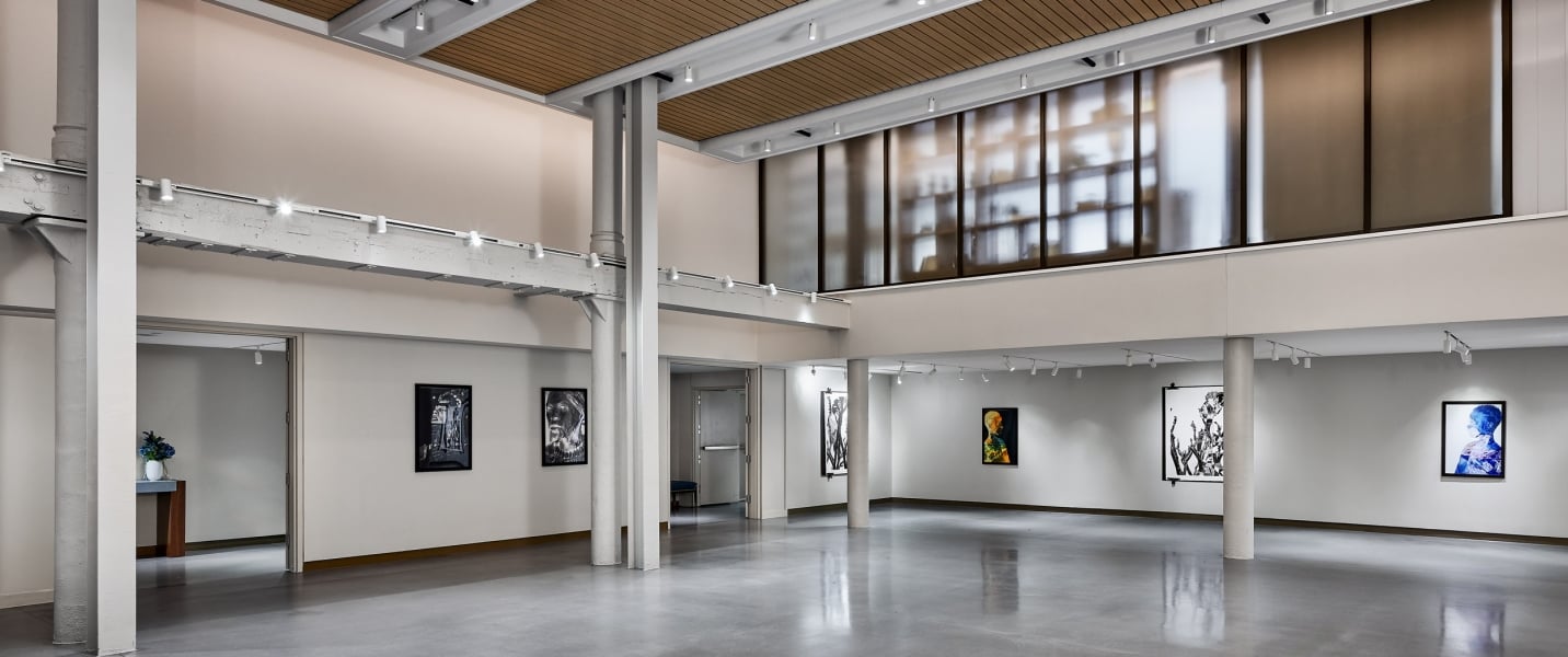 An empty modern art gallery displays various framed artworks on white walls, featuring a polished concrete floor, track lighting, and a slatted wood ceiling.