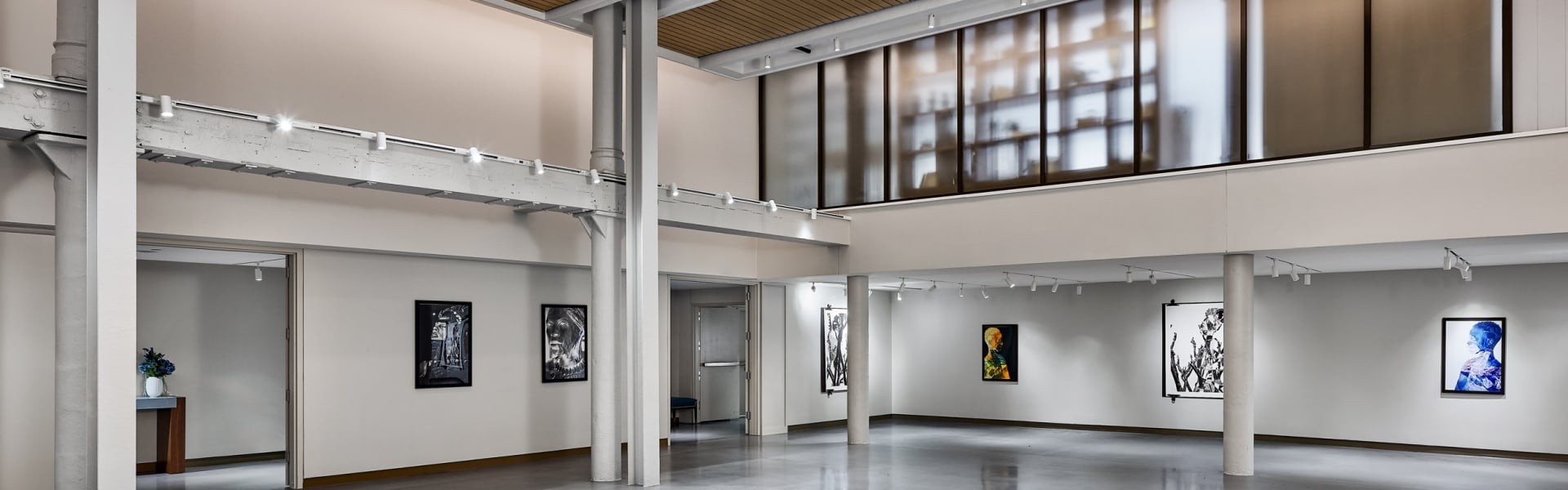 An empty modern art gallery displays various framed artworks on white walls, featuring a polished concrete floor, track lighting, and a slatted wood ceiling.