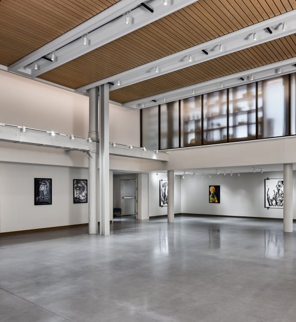 An empty modern art gallery displays various framed artworks on white walls, featuring a polished concrete floor, track lighting, and a slatted wood ceiling.