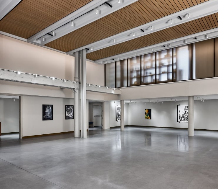 An empty modern art gallery displays various framed artworks on white walls, featuring a polished concrete floor, track lighting, and a slatted wood ceiling.