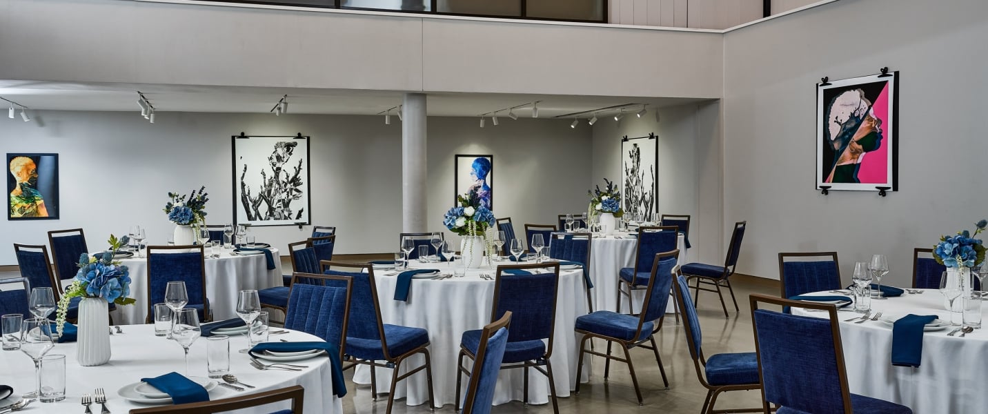 Round tables are set with white cloths, blue napkins, and floral centerpieces, surrounded by blue chairs. This modern event space features diverse artwork on light grey walls.