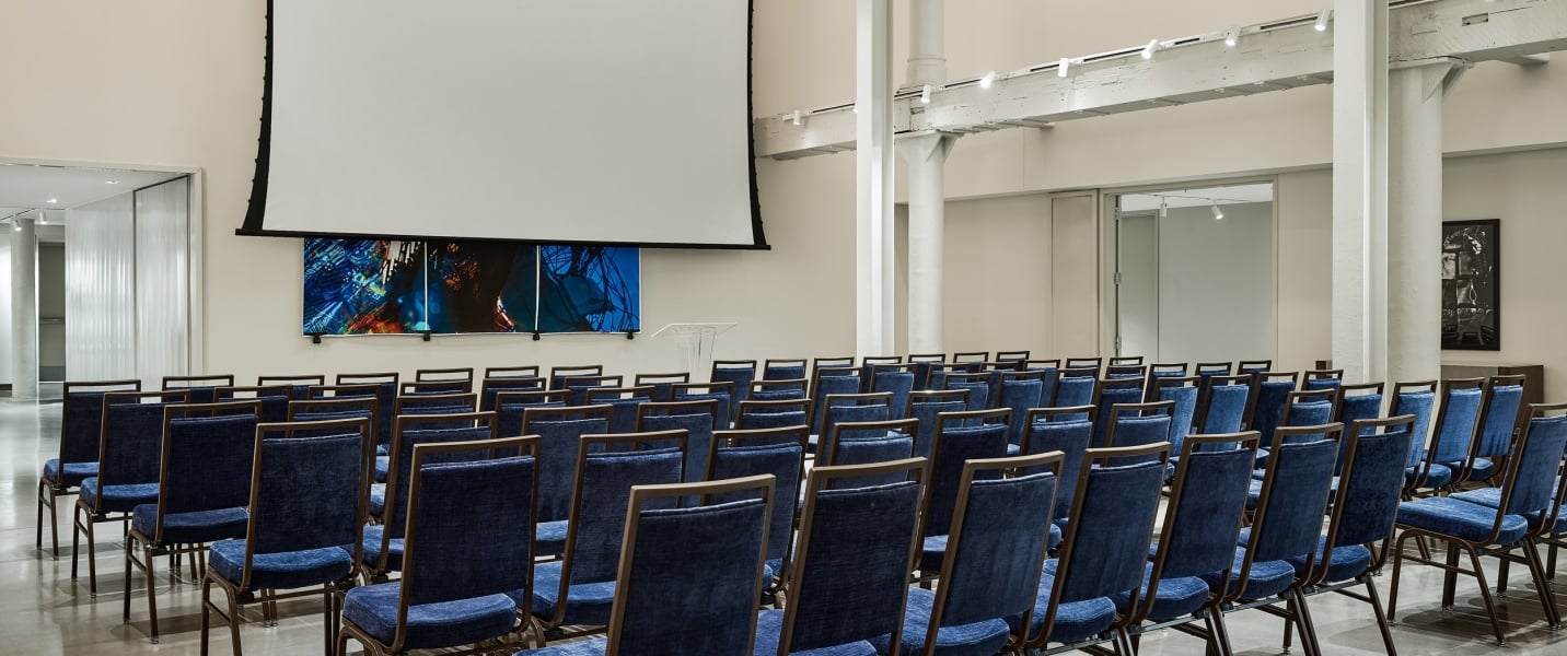 Blue chairs are neatly arranged in rows, facing a large projector screen and abstract art in a modern conference room.