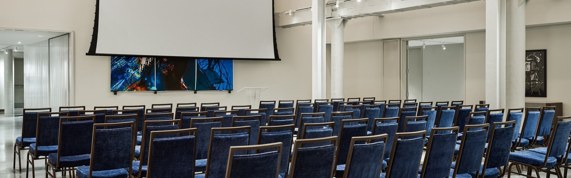 Blue chairs are neatly arranged in rows, facing a large projector screen and abstract art in a modern conference room.