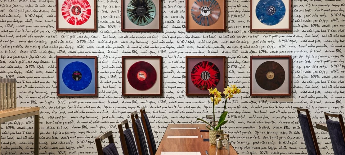 Nine framed, colorful vinyl records are displayed on a wall with black handwritten motivational phrases. A wooden table, chairs, and a yellow orchid are in the foreground.