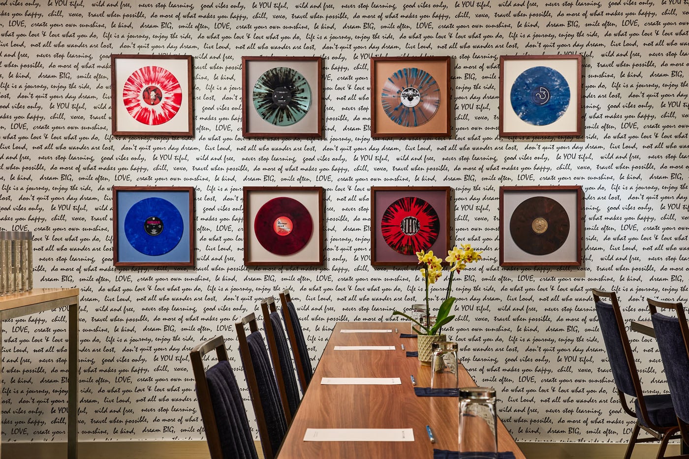 Nine framed, colorful vinyl records are displayed on a wall with black handwritten motivational phrases. A wooden table, chairs, and a yellow orchid are in the foreground.
