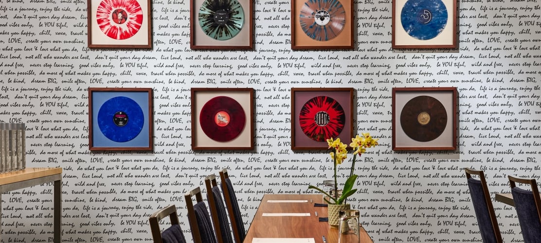 Framed vibrant vinyl records hang on a white wall covered in black handwritten quotes, above a long wooden table with chairs and a yellow orchid centerpiece.