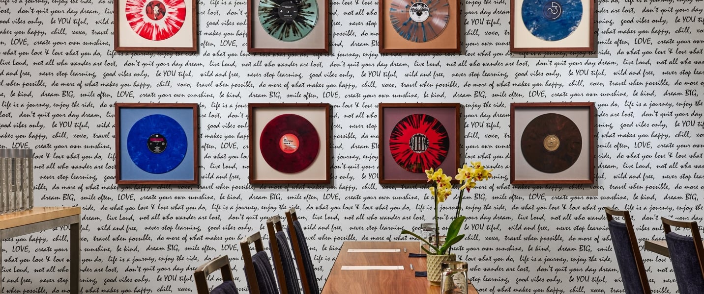 Framed vibrant vinyl records hang on a white wall covered in black handwritten quotes, above a long wooden table with chairs and a yellow orchid centerpiece.