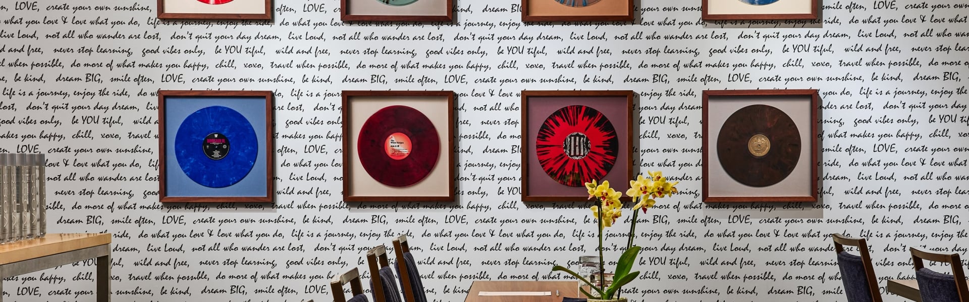 Framed vibrant vinyl records hang on a white wall covered in black handwritten quotes, above a long wooden table with chairs and a yellow orchid centerpiece.