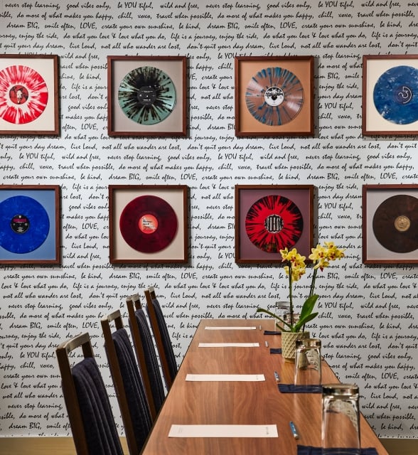 Framed vibrant vinyl records hang on a white wall covered in black handwritten quotes, above a long wooden table with chairs and a yellow orchid centerpiece.