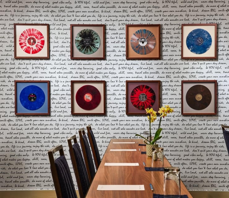 Framed vibrant vinyl records hang on a white wall covered in black handwritten quotes, above a long wooden table with chairs and a yellow orchid centerpiece.