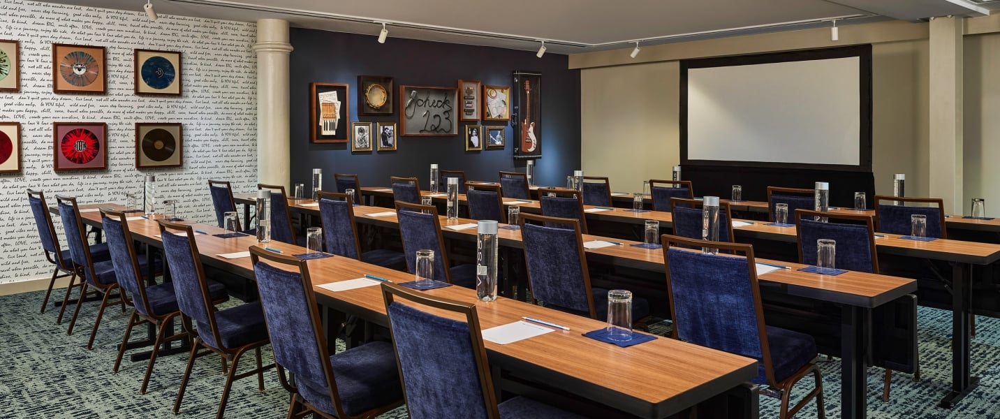 A conference room features rows of tables and blue chairs. Walls display music-themed decor, including framed records and a "Check 1 2 3" sign, opposite a projection screen.