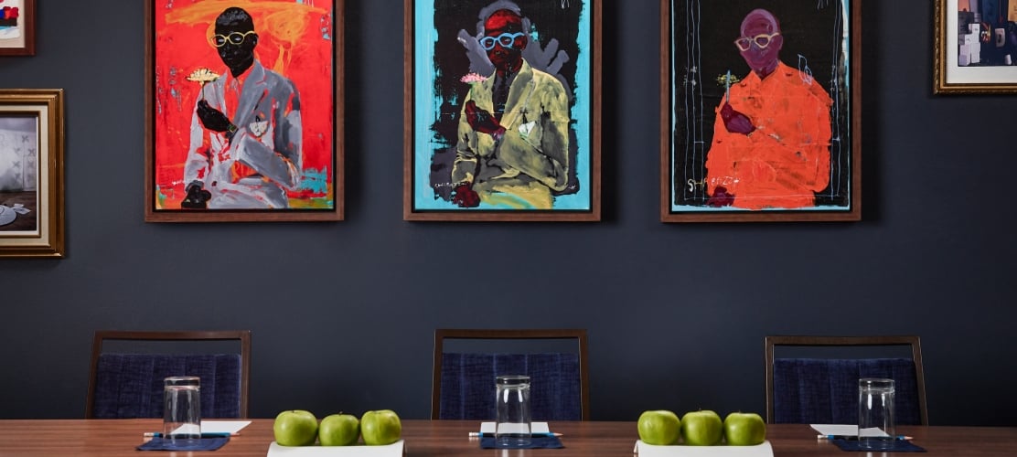 Three framed abstract paintings, signed SHABAZZ and depicting figures in suits, hang on a dark blue wall above several chairs.