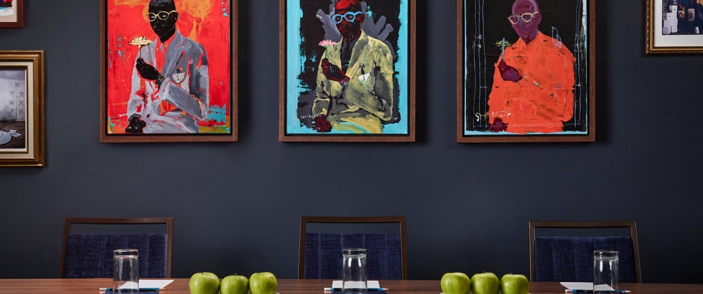 Three framed abstract paintings, signed SHABAZZ and depicting figures in suits, hang on a dark blue wall above several chairs.