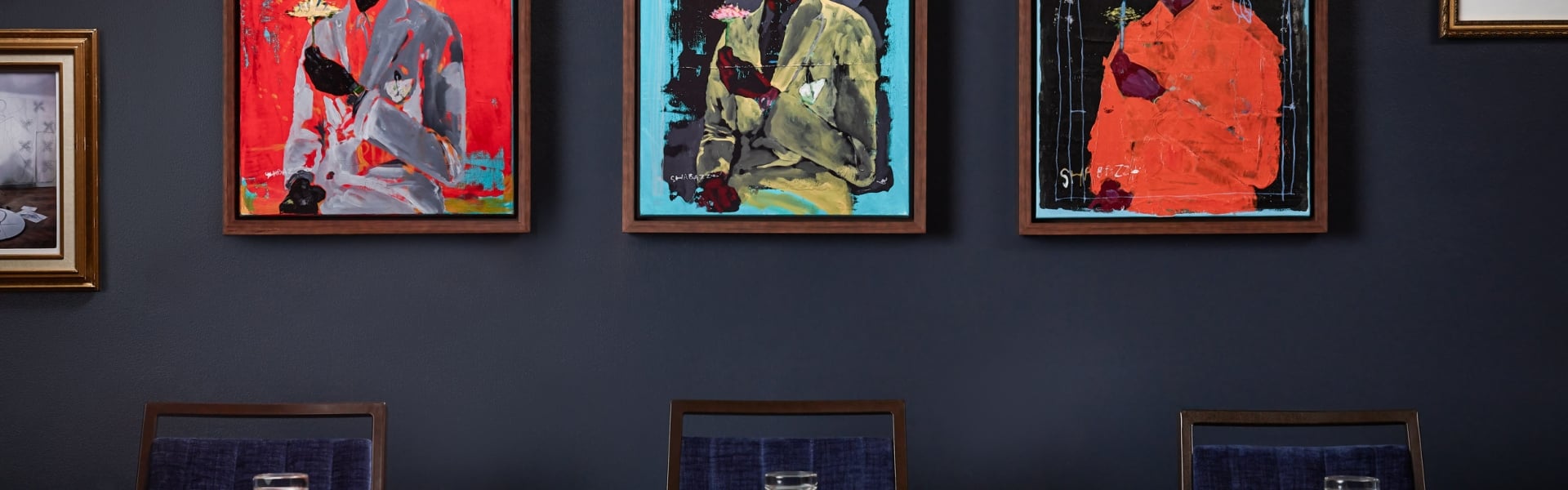 Three framed abstract paintings, signed SHABAZZ and depicting figures in suits, hang on a dark blue wall above several chairs.