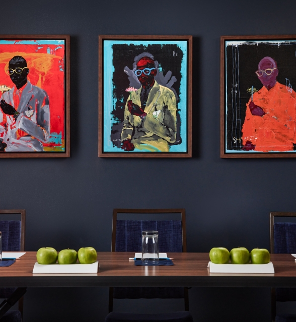 Three framed abstract paintings, signed SHABAZZ and depicting figures in suits, hang on a dark blue wall above several chairs.