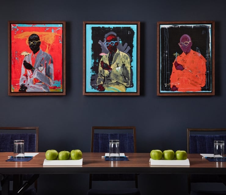 Three framed abstract paintings, signed SHABAZZ and depicting figures in suits, hang on a dark blue wall above several chairs.
