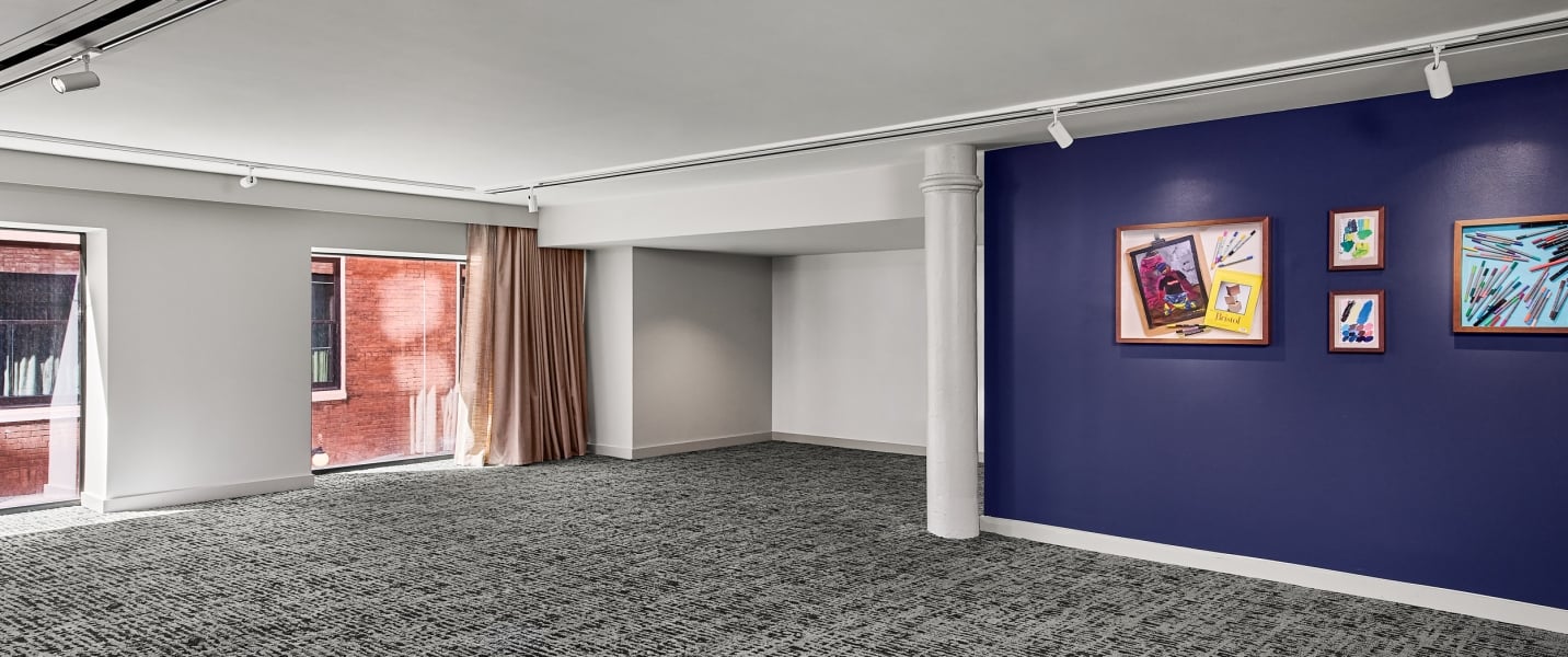 Children's artwork and art supplies are displayed on a navy blue wall in a spacious room with patterned gray carpet, large windows, and track lighting. Text: Bristol.
