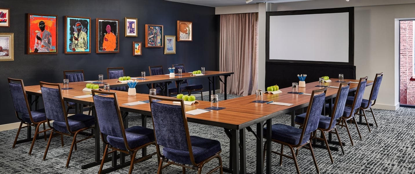 A U-shaped meeting table is set with chairs, water glasses, pens, and green apples. The modern room features colorful artwork on dark walls, a large projection screen, and patterned carpet.