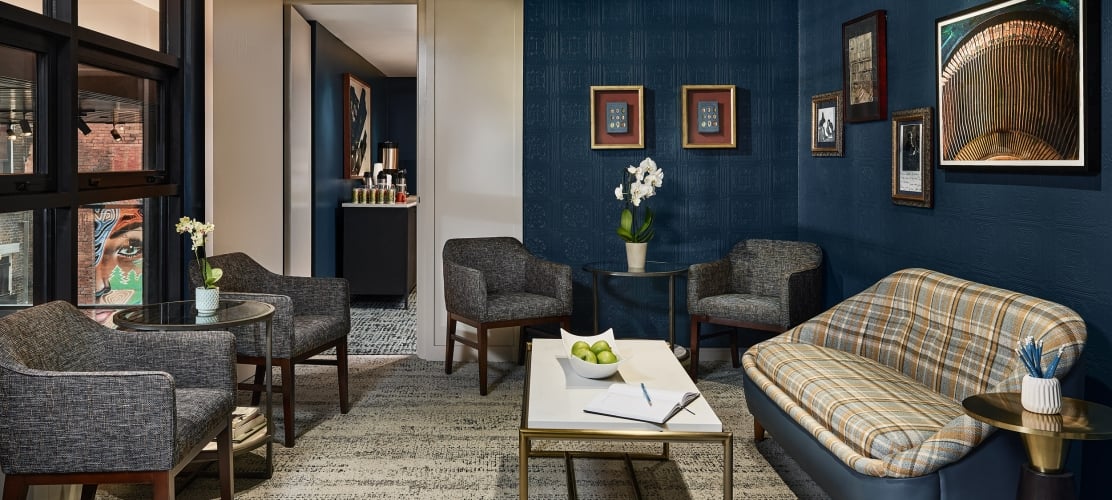 Stylish hotel lounge room featuring dark blue textured walls, framed art, a plaid sofa, and armchairs around tables with amenities, plus a visible beverage station.