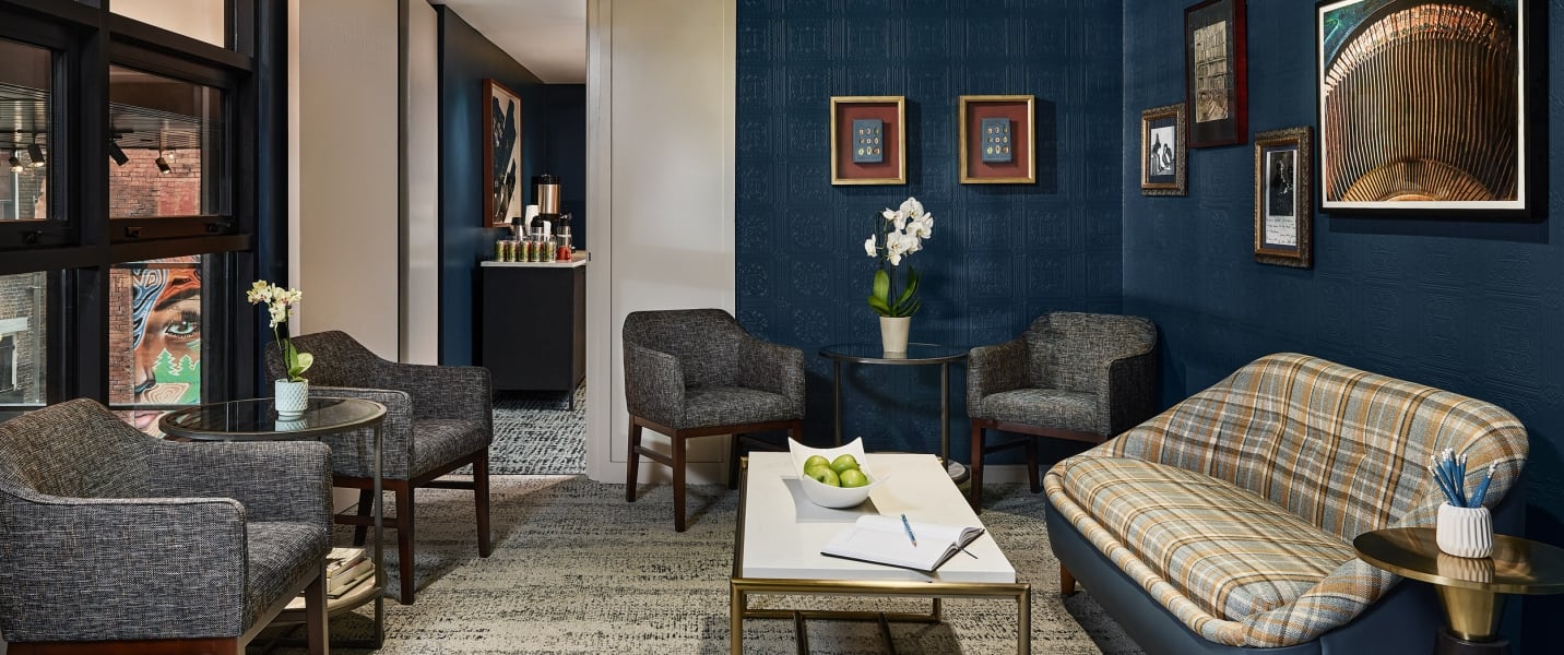 Stylish hotel lounge room featuring dark blue textured walls, framed art, a plaid sofa, and armchairs around tables with amenities, plus a visible beverage station.