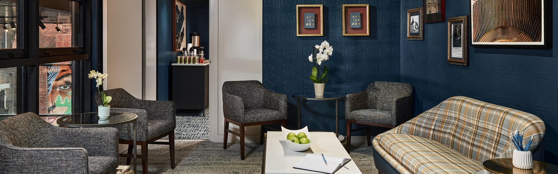 Stylish hotel lounge room featuring dark blue textured walls, framed art, a plaid sofa, and armchairs around tables with amenities, plus a visible beverage station.