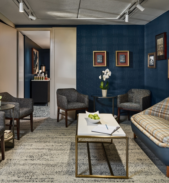 Stylish hotel lounge room featuring dark blue textured walls, framed art, a plaid sofa, and armchairs around tables with amenities, plus a visible beverage station.
