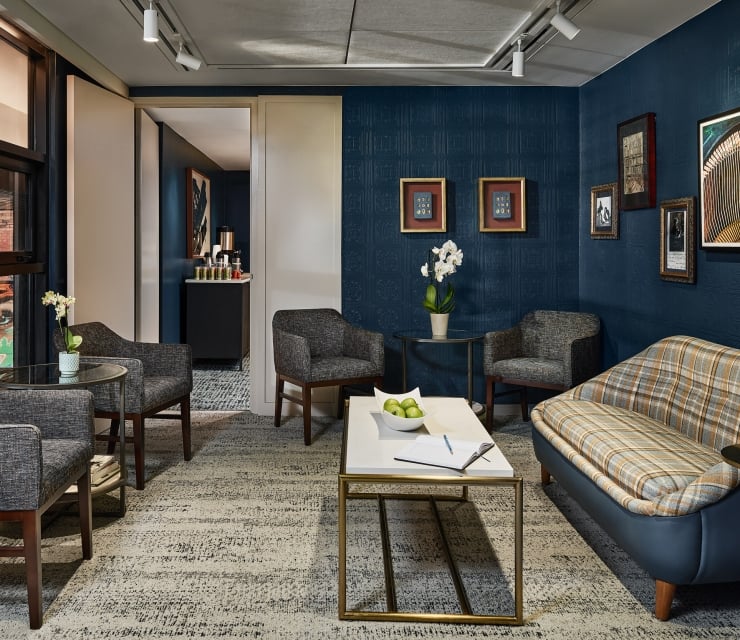 Stylish hotel lounge room featuring dark blue textured walls, framed art, a plaid sofa, and armchairs around tables with amenities, plus a visible beverage station.