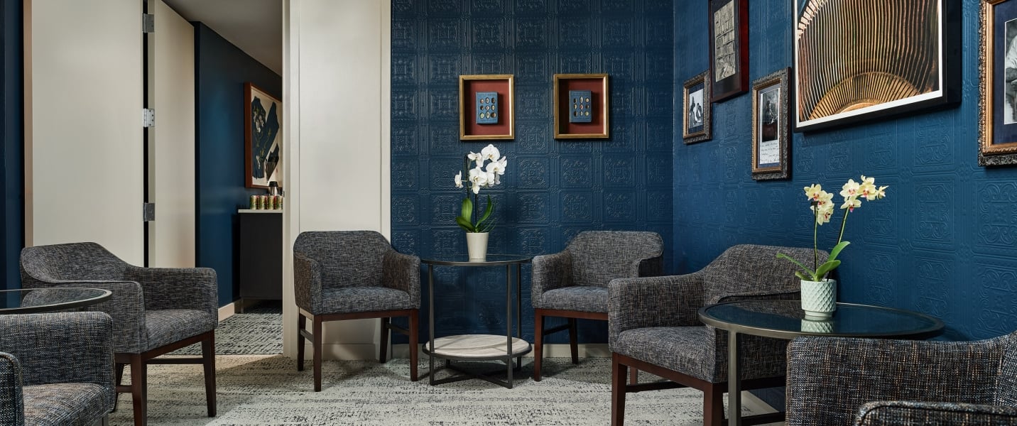 Upholstered armchairs and small tables are arranged on a patterned rug in a room with deep blue embossed walls, framed artwork, and white orchids.