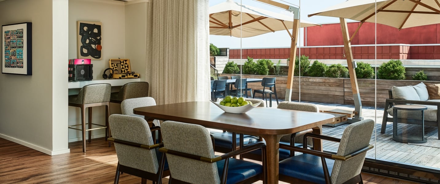 A wooden dining table with green apples sits in a modern indoor-outdoor room. It connects to an outdoor patio with umbrellas, seating, and planters.