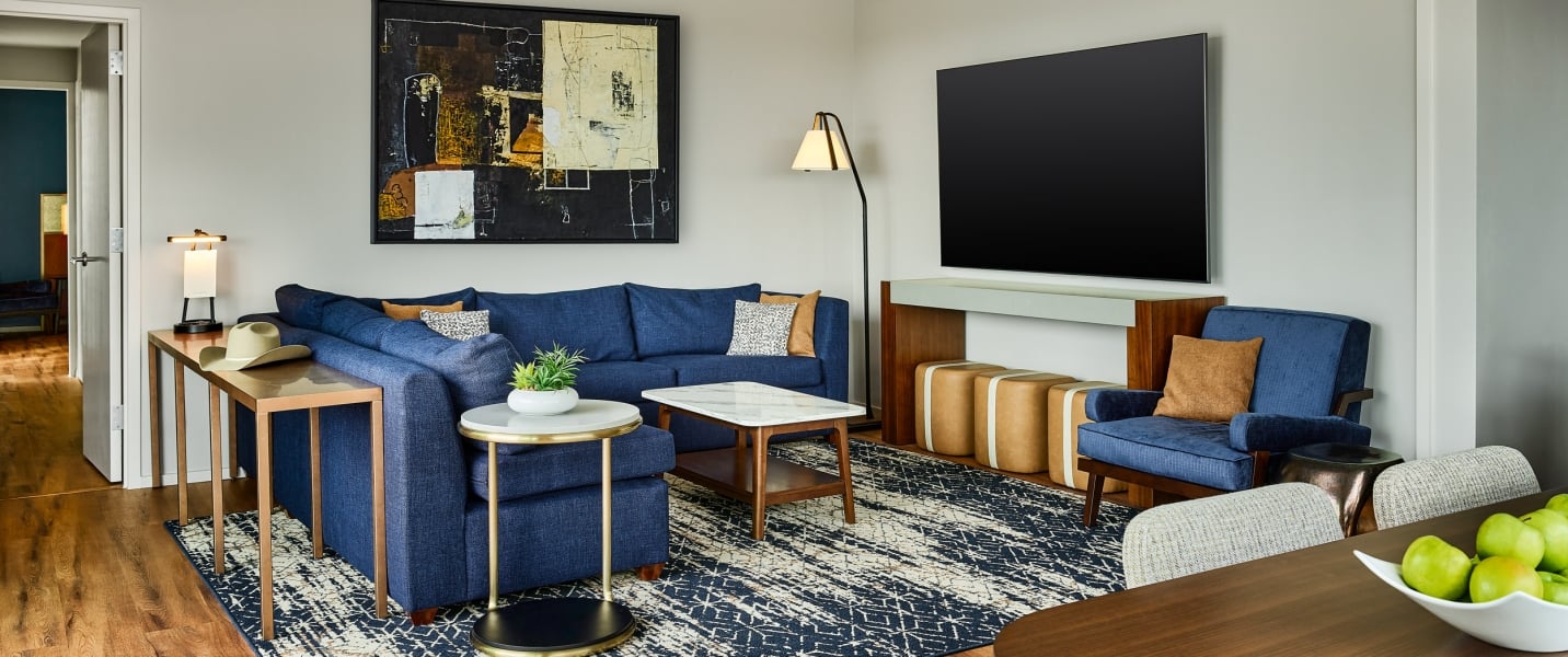 A blue sectional sofa and armchair are arranged with a coffee table, console holding a cowboy hat, and an abstract painting in a contemporary living room with a large TV.