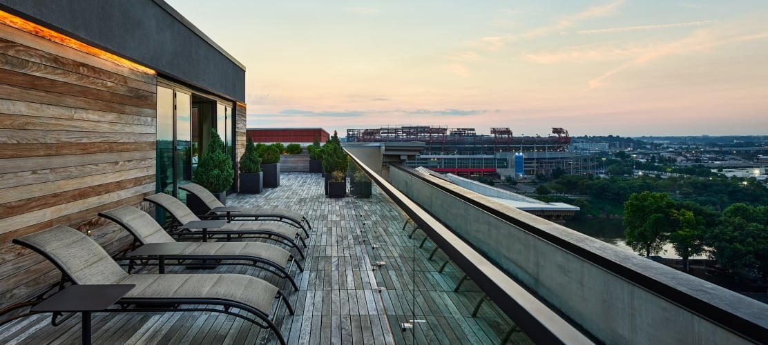 Several lounge chairs line a wooden rooftop deck, overlooking a city stadium and a river under a sunset sky.