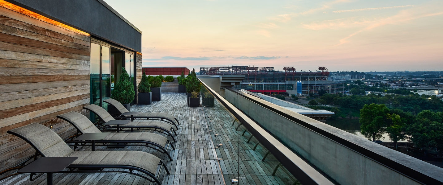 Several lounge chairs line a wooden rooftop deck, overlooking a city stadium and a river under a sunset sky.