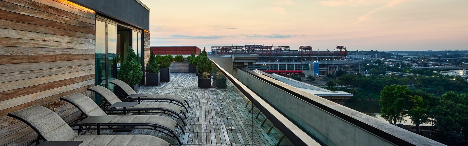 Several lounge chairs line a wooden rooftop deck, overlooking a city stadium and a river under a sunset sky.