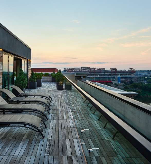 Several lounge chairs line a wooden rooftop deck, overlooking a city stadium and a river under a sunset sky.