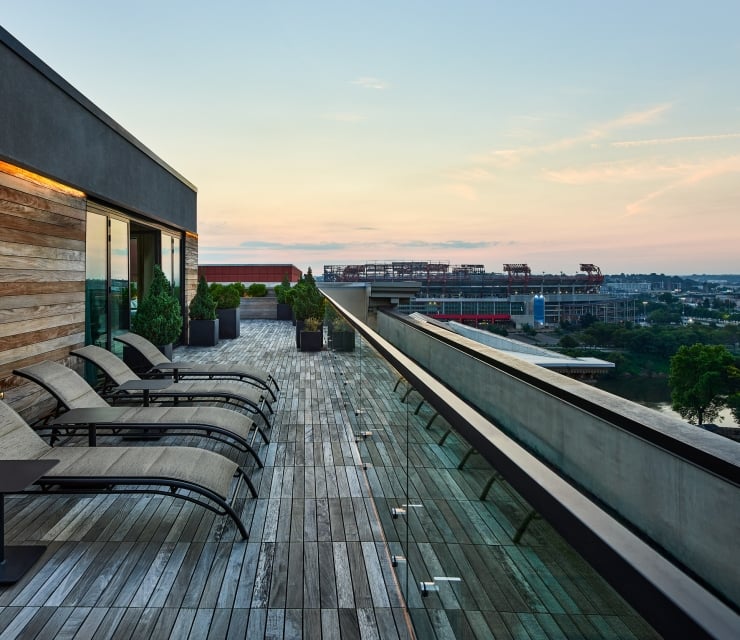 Several lounge chairs line a wooden rooftop deck, overlooking a city stadium and a river under a sunset sky.