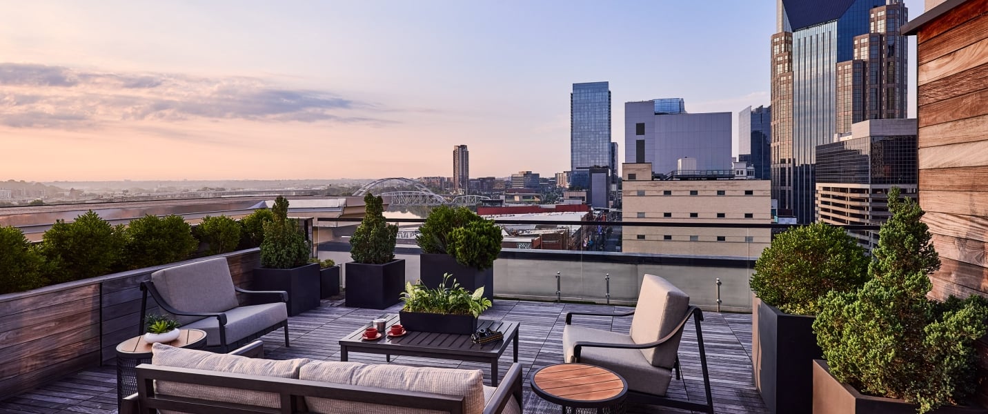 A spacious rooftop patio features outdoor seating on wooden decking, with planters bordering the space. It overlooks a city skyline with skyscrapers and a bridge under a soft, sunset sky.