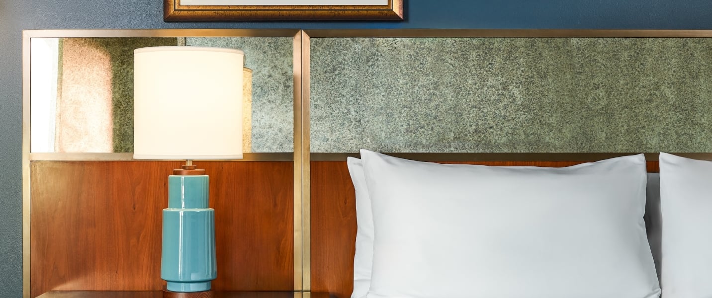 An illuminated teal table lamp stands on a nightstand next to white pillows on a bed. It's set against a wood and speckled headboard under a blue wall.