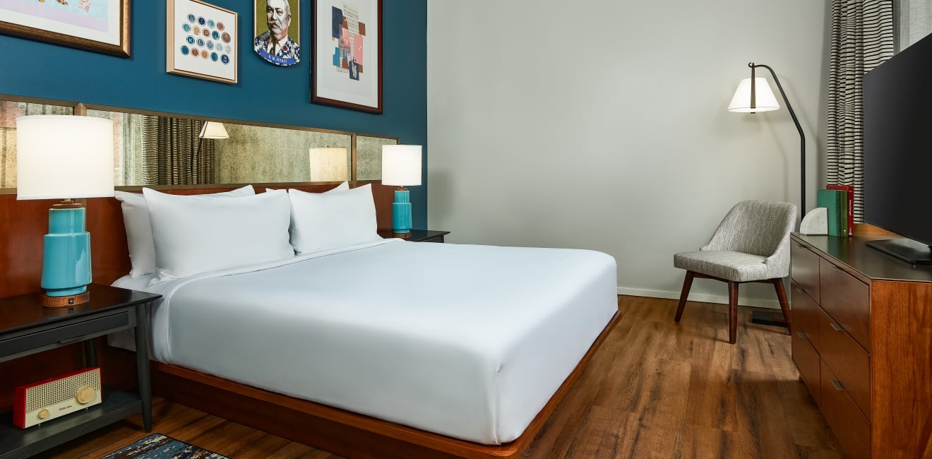 Modern hotel bedroom featuring a king bed with a dark wood headboard, a blue gallery wall with framed art, a mid-century dresser, and a grey armchair in the corner.