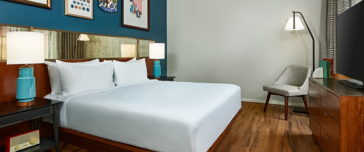 Modern hotel bedroom featuring a king bed with a dark wood headboard, a blue gallery wall with framed art, a mid-century dresser, and a grey armchair in the corner.
