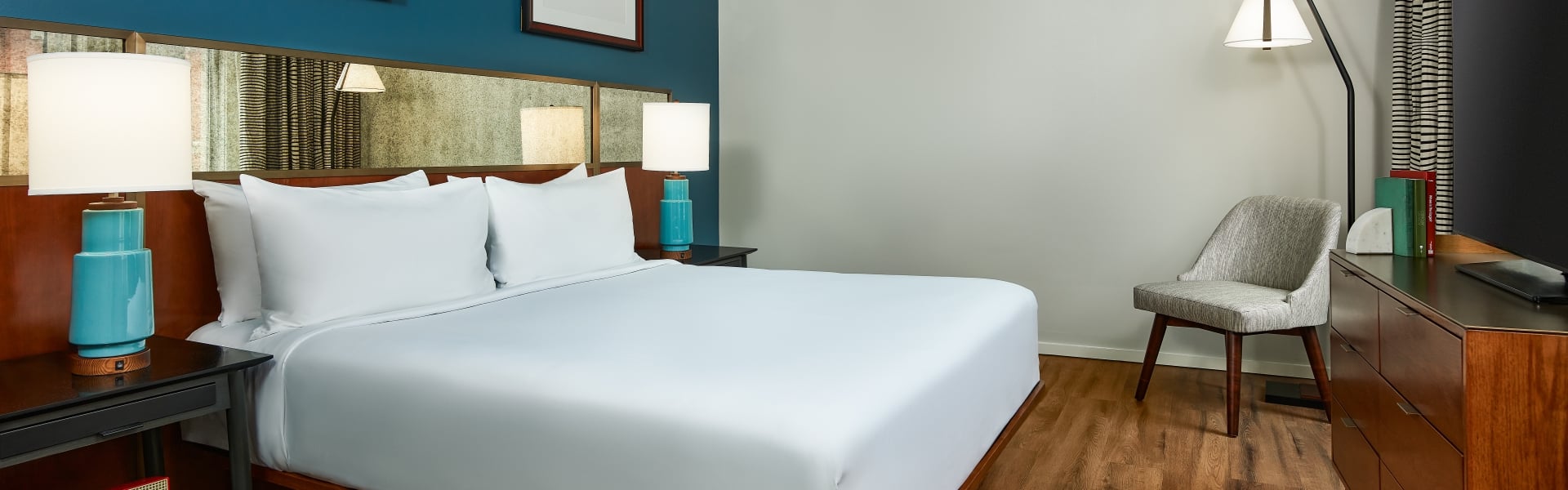 Modern hotel bedroom featuring a king bed with a dark wood headboard, a blue gallery wall with framed art, a mid-century dresser, and a grey armchair in the corner.