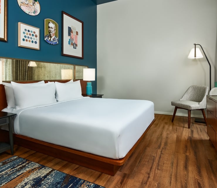 Modern hotel bedroom featuring a king bed with a dark wood headboard, a blue gallery wall with framed art, a mid-century dresser, and a grey armchair in the corner.