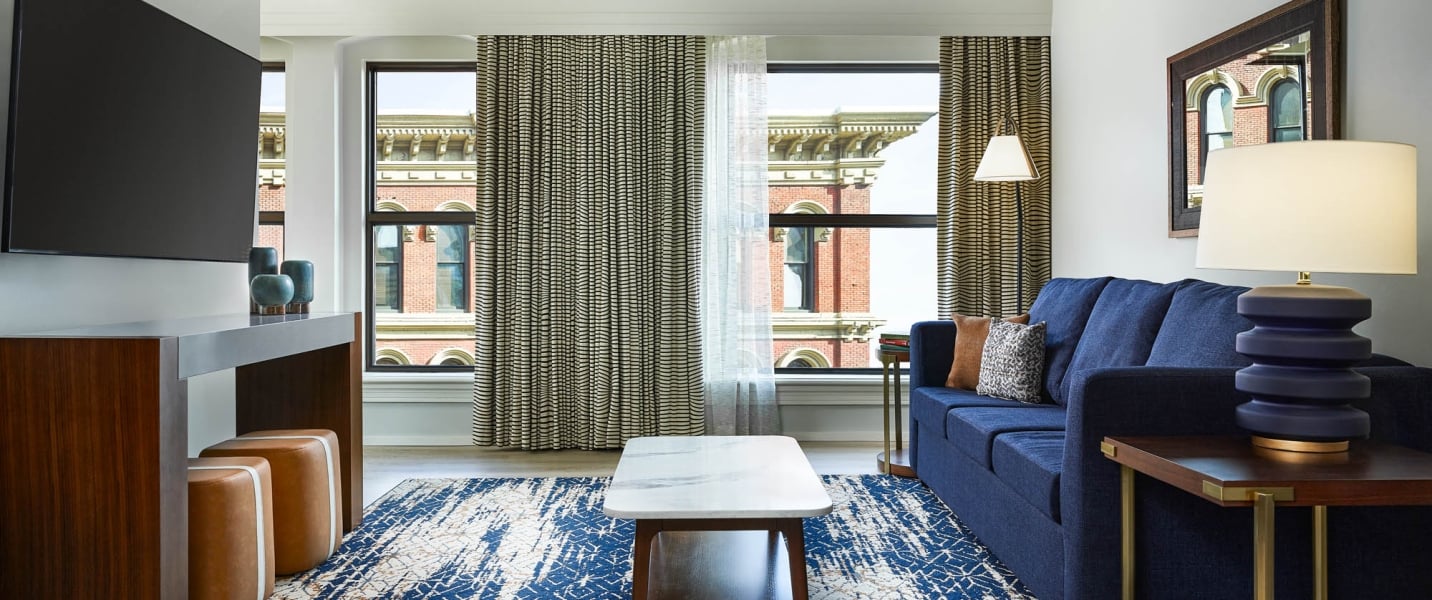 A modern living room features a blue sofa, TV, and patterned rug opposite large windows showing an ornate brick building.
