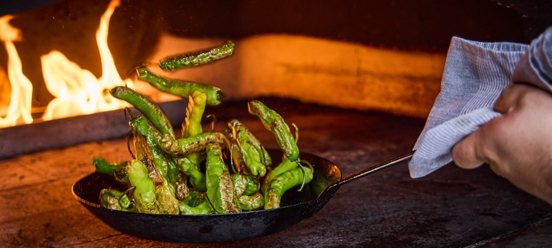 A person tossing shishito peppers in a pan over an open flame oven