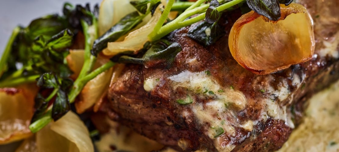 Grilled meat topped with a creamy herb sauce, garnished with vibrant green leaves, roasted onion petals, and golden potato chunks, presented as a gourmet meal.