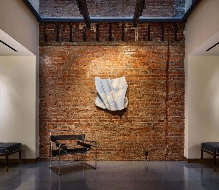 An art gallery wall with an exposed brick center section featuring a draped, light-blue textile sculpture. Modern seating benches and additional artwork frame the installation under a skylight.