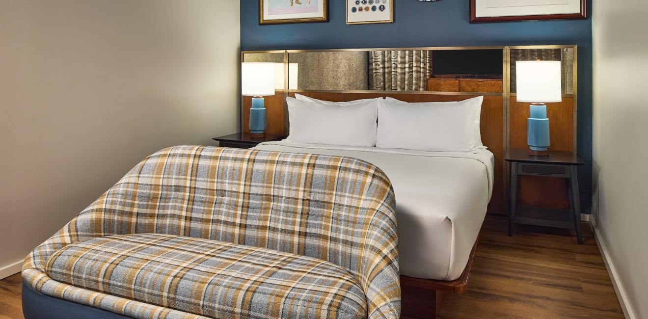 A modern bedroom features a bed with white linens and two lamps, facing a plaid bench. Blue walls with art hang over a wooden floor.