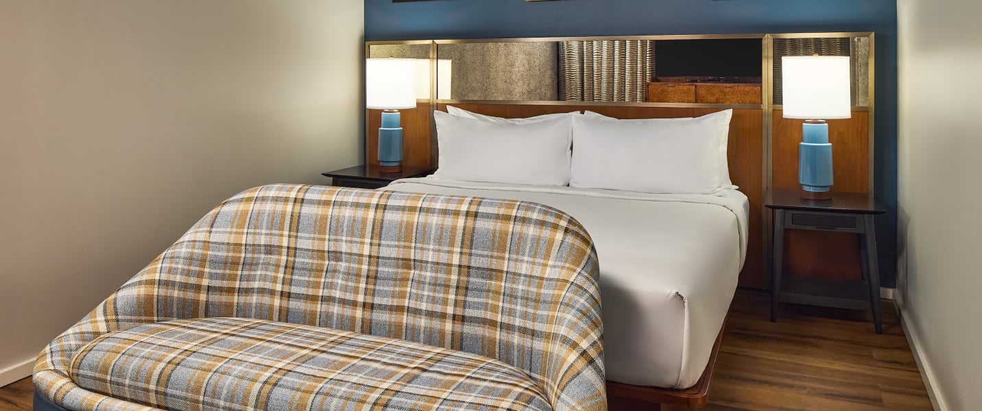 A modern bedroom features a bed with white linens and two lamps, facing a plaid bench. Blue walls with art hang over a wooden floor.