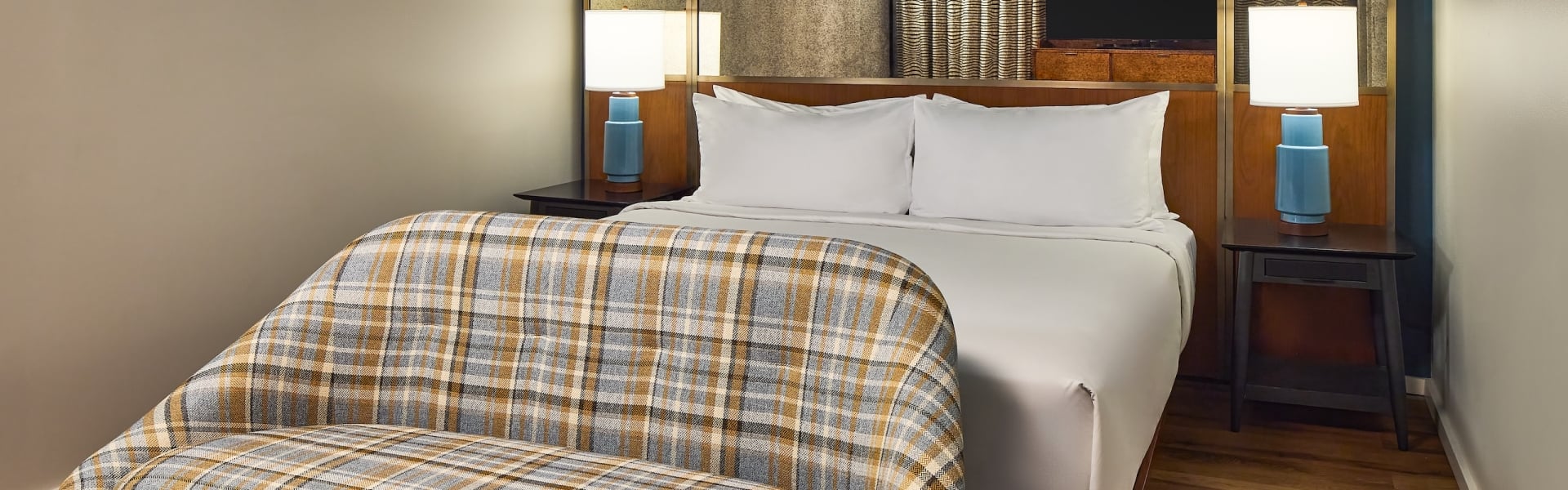 A modern bedroom features a bed with white linens and two lamps, facing a plaid bench. Blue walls with art hang over a wooden floor.
