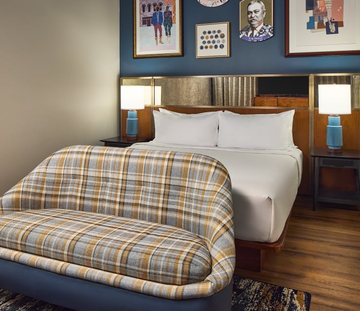 A modern bedroom features a bed with white linens and two lamps, facing a plaid bench. Blue walls with art hang over a wooden floor.