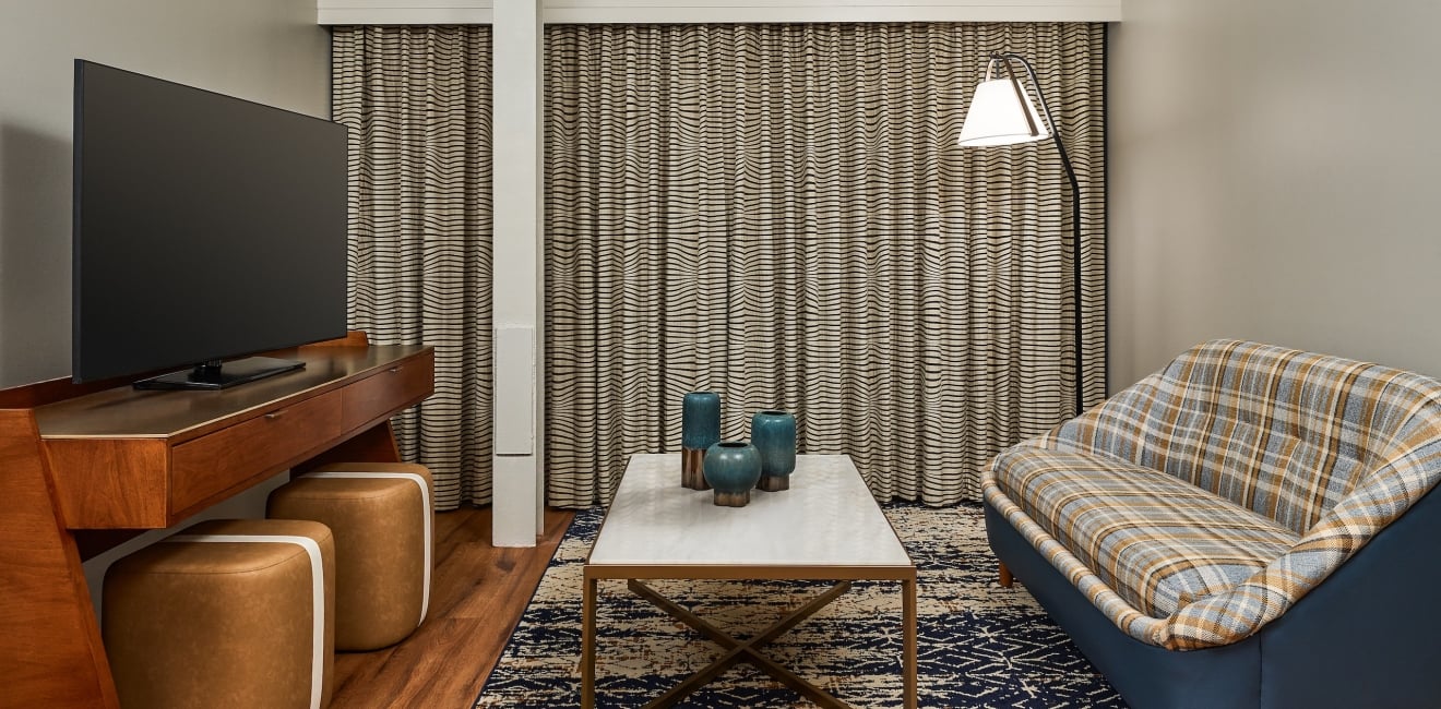 Modern living area featuring a large TV, a coffee table with three blue vases, and a plaid sofa. Wavy-striped curtains cover the back wall.